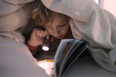 menino de bruços segurando uma lanterna para ler um livro dentro de uma cabana de lençol